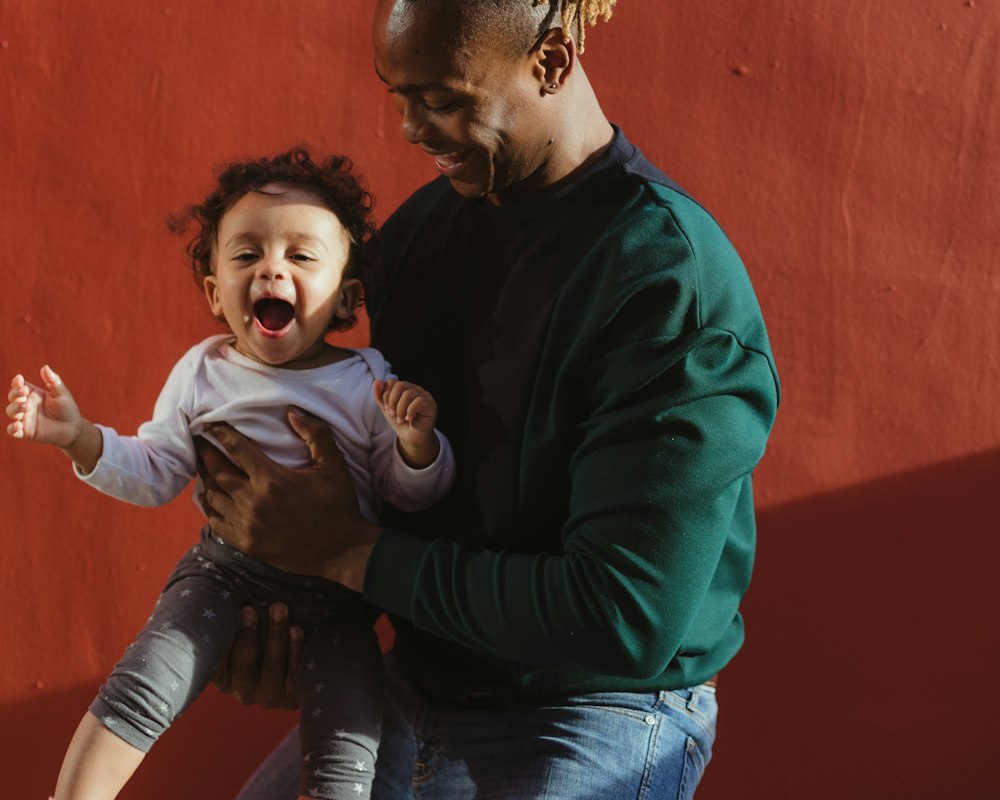 A smiling dad holding his laughing baby against a red wall
