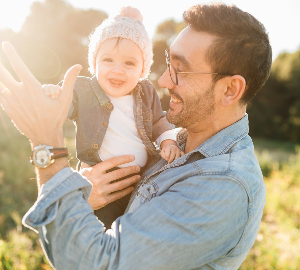 A smiling dad in a denim jacket holding his baby in warm evening light